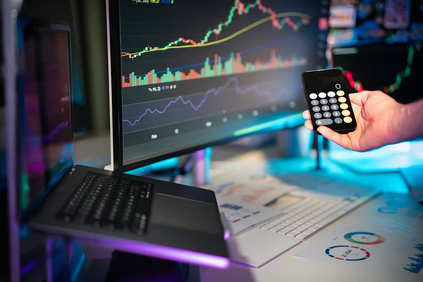 Ruka drží telefón s kalkulačkou pri finančných údajoch na monitoroch.